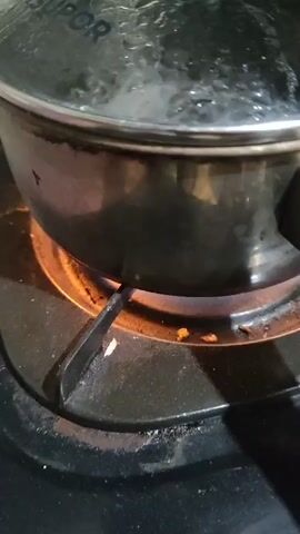 A close-up shot captures a black pot with a clear lid sitting on a gas stove burner. The burner is lit, casting an orange glow around the base of the pot. Inside the pot, water is visibly boiling, creating bubbles and steam that rise to the underside of the lid. The lid has some condensation on it, and a faint brand name "SUPOR" is visible at the top. As the video progresses, the lid is removed, revealing more vigorously boiling water and steam escaping the pot. The background is dark, with hints of a green surface visible in the upper portion of the frame.