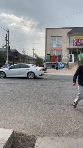 The video shows a street scene in Ismoili Somoni, Tajikistan, on a cloudy day. A white sedan is parked on the left side of the road, facing away from the camera. Across the street is a two-story building with a light-colored facade. The ground floor of the building appears to house several shops, indicated by the signage and displays. One prominent sign reads "МАГОЗАИ ОИЛАВИ" (Family Store) above a graphic featuring four people. Below this, the word "ORO" is visible, likely the name of a shop.

A person is walking in front of the building. A dark-colored sedan is driving past the building from left to right. Another dark-colored sedan is parked in front of the building. The overall atmosphere is that of a typical street in a town, with shops open and people going about their business. The cloudy sky suggests it might be late afternoon or early evening. The presence of traffic lights on a pole to the left of the frame indicates this is likely an intersection.
