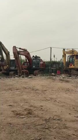 The video shows an outdoor area with construction equipment and two people. In the foreground, there is a dirt and gravel ground. In the background, several excavators are parked. A red excavator is prominent in the center, with a yellow excavator to its right. To the left of the red excavator, a portion of another excavator is visible. Further back, a wooden guard tower with a red roof stands on stilts. There is a fence behind the guard tower, and some greenery and trees are visible in the distance. Two individuals are walking away from the camera, towards the right side of the frame. One person is wearing an orange shirt and dark pants, and the other is wearing a white shirt and dark pants. The sky is overcast.