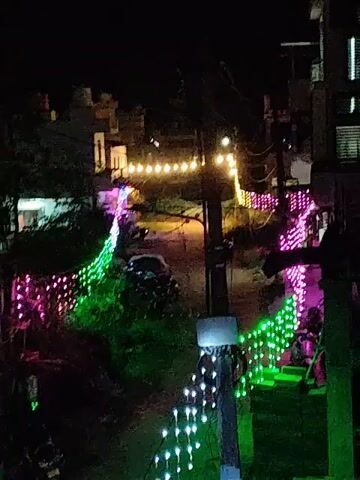 The video shows a nighttime view of a street decorated with colorful lights. The lights are strung along wires, illuminating the street and the sides of buildings. There are also lights in the distance, suggesting a broader festive atmosphere. The scene appears to be in a residential area with houses and possibly some shops visible. A few vehicles, including a car and motorcycles, are parked or driving on the street. The lighting creates a vibrant and festive ambiance.