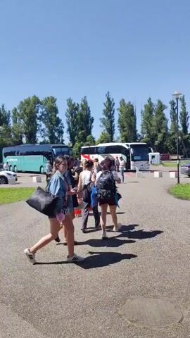 A sunny day with a clear blue sky. The video shows a parking lot with several tour buses and cars. People are seen walking around, some carrying bags, suggesting they are travelers. Trees line the background, and streetlights are visible. The focus shifts as the camera moves, capturing different angles of the scene.