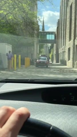The view from inside a parked car shows a narrow street framed by stone buildings on either side. Sunlight peeks through tree branches in the upper left corner. A red SUV is parked a short distance ahead, with two people interacting near the driver's side door. A person wearing a purple shirt walks near yellow poles on the left sidewalk. The sky is blue with some clouds. A covered walkway connects the two buildings, and a church steeple is visible in the background. The dashboard and steering wheel of the car are visible in the foreground, showing the car is running.