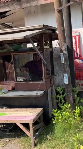A woman wearing a purple headscarf and maroon shirt is standing behind a food stall. She is looking to her left and holding a cooking utensil in her right hand over a frying pan. The food stall is wooden and appears weathered. It has shelves with various items, including green bowls, plastic bags, and a blue basket containing fried food. A pink and green painted wooden bench is in front of the stall. The background shows a building with a tiled roof and a concrete wall. Sunlight suggests it is daytime.