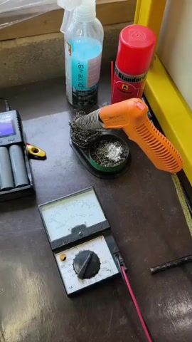 The video shows a workbench with various electronic testing and charging equipment. In the foreground, an analog multimeter with red and black leads is visible. To the left, a digital battery charger with four AA batteries is plugged in and displaying information on its screen. Next to the charger, an orange sits in a black plastic holder. Behind these items, a spool of solder and a soldering iron with a metal brush cleaning pad are present. To the right of the soldering iron, a red can of spray and a clear spray bottle with blue liquid are on the bench. The background appears to be a wooden surface with some plastic sheeting. The lighting suggests an indoor environment.