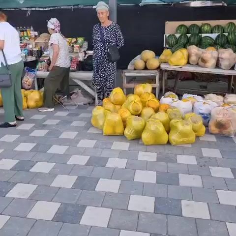 A busy outdoor market scene unfolds with vendors and customers interacting around stalls filled with produce. Several people are visible, including women in headscarves and men in casual attire. Large piles of yellow-bagged produce, possibly melons or citrus fruits, are prominent in the foreground. Behind these, tables display watermelons, pumpkins, and other fruits and vegetables. The ground is paved with a checkerboard pattern of gray and white tiles. Some individuals are holding shopping bags, and the overall atmosphere suggests a typical market day with transactions and movement.