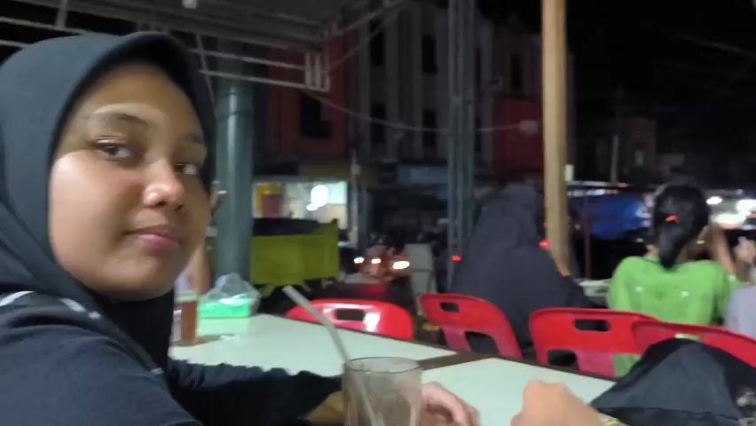 A group of people are seated at a long table with red chairs, enjoying drinks at an outdoor cafe or restaurant at night. The woman at the beginning of the video is wearing a black hijab and is holding a glass. The camera pans to show a woman in a blue hijab using her smartphone. There are other people seated around the table, some with their backs to the camera, wearing casual clothing.  The setting appears to be a bustling street in an urban area, with buildings and vehicles visible in the background. The atmosphere seems relaxed and social.