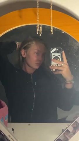 A young person with blonde hair takes a mirror selfie indoors. They are wearing a dark, zip-up jacket. In the first part of the clip, their hair is loose, and they adjust it with their left hand, looking slightly to the side with a neutral expression. In the second part, they rest their left hand on their chin, looking directly at their reflection with a more thoughtful or pensive look. Their right hand holds a light-colored smartphone with a sticker featuring a circular design on the back. They are wearing multiple rings on their right hand and bracelets, including one with shell-like beads, on their right wrist.

The mirror is round with a prominent light brown or orange wooden frame. Several delicate gold-colored necklaces hang from above the mirror, reflecting in its surface. The mirror itself appears somewhat smudged. The background reflected in the mirror is dark and indistinct, while the wall around the mirror is a light, possibly off-white, color. Below the mirror, a pink item (possibly a bag) is visible on the left, and some papers or photos are on the right, one showing partially visible text like "motion me." The setting appears to be a personal indoor space, possibly a bedroom, with dim lighting. No specific location cues for Lannoy, France are visible.