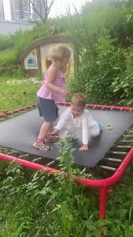 Two children are playing on a rectangular trampoline. The trampoline has a red frame and a black mat. One child, a boy, is in a crawling position on the trampoline. He is wearing a white long-sleeved shirt and blue shoes. The other child, a girl, is standing and appears to be jumping. She is wearing a pink sleeveless top and dark shorts. Behind the children is a small, whimsical structure resembling a hobbit house with a round door. The area is surrounded by lush green grass and vegetation, with tall buildings visible in the background. The sky is overcast.