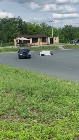 A black Subaru WRX hatchback performs a drift on an asphalt track, kicking up dust and smoke. A person wearing a grey baseball cap and black t-shirt stands in the foreground, filming the car with a smartphone. In the background, a few other cars are parked, along with some buildings and trees. The sky is cloudy. The video is shot from a low angle, with the grass and a concrete barrier in the foreground.