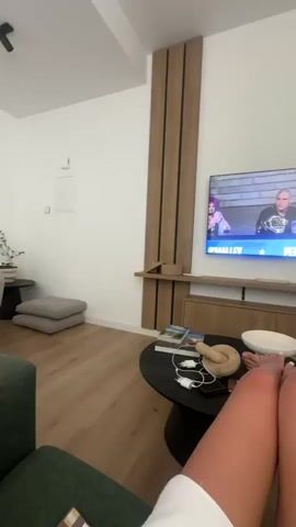 The video shows a first-person perspective of someone relaxing in a living room, watching a television. The television displays a broadcast with three people sitting at a table, and two championship belts are visible. In the foreground, the legs and feet of two individuals are prominent, suggesting they are either sitting or lying down together. A coffee table in front of them holds a bowl, some magazines, and what appears to be a toy or plush item. The room has modern wooden paneling on the wall behind the TV and light-colored hardwood floors. To the left, a portion of a grey sofa is visible, and to the right, an armchair with a patterned cushion is present. The overall ambiance is one of leisure and relaxation.