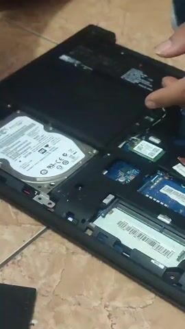 Here is a concise description of the video:

A person is unscrewing a hard drive from the open back of a laptop using a screwdriver. The laptop is black and resting on a tile surface. You can see other components inside the laptop as well, including what appears to be memory and wireless network cards. There are a few loose screws nearby.