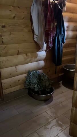 The video shows the interior of a sauna or bathhouse. The walls are made of horizontal wooden logs. On the wall, there are towels and a robe hanging on hooks. At the base of the wall, there is a metal basin containing a bunch of leafy branches. The floor is tiled with light brown rectangular tiles. Some water is visible on the floor. The footage is slightly shaky and the lighting is dim. The camera pans upwards along the wall.  A curtain or sheet is seen hanging in the scene. The overall impression is a rustic and cozy atmosphere. Audio of a person speaking is present.