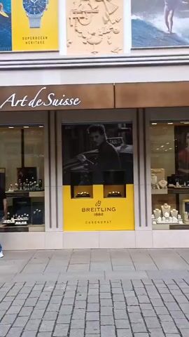 The video pans across a cobblestone street lined with shops and historic buildings. On the left, a man walks away from the camera in front of a watch shop called "Art de Suisse." The shop window displays various watches and jewelry. Further down the street, a display of clothing can be seen through the window of another shop. On the right side of the street, a large yellow sign with "BREITLING" is visible on a building. Two young men walk towards the camera. Another shop, "Nareis," displays clothing on mannequins. A woman with blonde hair and sunglasses walks towards the camera, followed by another woman. A building with red brick and ornate architectural details stands prominently. The street is clean and well-maintained, suggesting a commercial or tourist area. The overall atmosphere appears to be a pleasant, sunny day.
