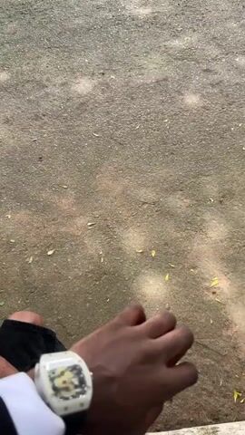 The video shows a person's point of view while sitting outdoors. They are wearing a watch with a silver case and a black band on their left wrist. The ground is asphalt with some leaves scattered on it. Other people are walking around. In the background, there's a grassy field with trees and some buildings.