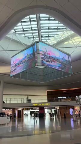 The video pans across a brightly lit, modern airport terminal. The ceiling is a high, vaulted structure with a large skylight revealing a network of beams. Suspended from the ceiling is a large, multi-faceted LED screen displaying colorful, abstract animated graphics. Below the screen, on the ground floor, there is a wide, polished floor reflecting the overhead lights. In the background, people can be seen walking and standing around. There are shops and signs visible on a higher level. The overall impression is one of a busy, contemporary transportation hub.