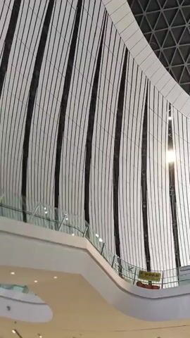 The camera pans upwards to display the interior of a building with a unique ceiling design. The lower portion of the frame showcases vertical, light-colored panels interspersed with dark, narrow stripes, complemented by a curved, white balcony. The upper portion of the frame is dominated by a complex geometric structure, a dome-like grid of interconnected triangles in a dark shade, potentially black or gray. The lighting seems to be a mixture of natural light filtering through the structure and artificial lights embedded within the panels. The overall environment gives the impression of a modern, spacious building, possibly an airport, shopping mall, or convention center. There are no people clearly visible in the frame. The focus is on the architectural design of the interior space.