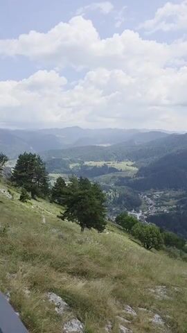 The video shows a panoramic view of a mountainous landscape on a cloudy day. In the foreground, there is a grassy slope with a few scattered trees and rocks. A metal railing runs along the edge of the slope, providing a safety barrier. The railing is made of metal bars and a thicker top rail. Beyond the railing, the view opens up to reveal rolling hills and valleys covered in dense green forests. In the distance, more layers of mountains are visible, fading into a hazy blue. A small village or settlement with some buildings and a church steeple can be seen nestled in one of the valleys. The sky is a mix of blue and white clouds, with patches of sunlight breaking through. The camera slowly pans across the landscape, capturing the vastness and natural beauty of the scene. There are no people visible in the video.