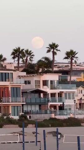 The video shows a full moon rising above a row of modern multi-story beach houses with palm trees in front. A volleyball net is visible in the foreground on a sandy beach. The sky is a pale, hazy pink, indicating either dawn or dusk. The houses are light-colored with large windows and balconies. A single person is visible walking on a balcony on the left side of the frame.