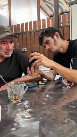 A man wearing a camouflage cap looks at a phone that another man, wearing a black t-shirt, is holding. They are sitting at a table with a transparent plastic tablecloth. A teacup with some liquid and a spoon is on the table in front of the man with the cap. Other objects, possibly tools, are also visible on the table. The background appears to be an outdoor or semi-outdoor setting with wooden walls and a corrugated metal roof. The younger man is holding the phone in his hands and appears to be showing something on the screen to the older man. The older man is looking intently at the phone, with a serious expression.