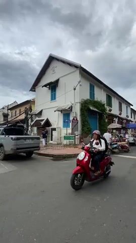 A red scooter driven by a person wearing a white long-sleeved shirt, blue pants, and a pink hat with a black backpack rides past a white building with blue shutters. The scooter is in the foreground, slightly to the left of the frame. The building is a two-story structure with a peaked roof and a green vine growing up its side. Several other scooters are parked in front of the building. To the left of the building, a silver SUV is parked. The sky is overcast and cloudy. The street is paved and appears to be in a city or town.