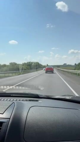 A first-person perspective video shows a car driving on a highway on a sunny day. The view is from the driver's seat, looking forward through the windshield. The dashboard of the car is visible in the foreground. Two cars are ahead on the highway, one red and one white. The red car is further ahead and closer to the center of the frame. The white car is to the left and further in the distance. The highway is paved and has multiple lanes. Guardrails are visible on the sides of the road. Trees and vegetation line the sides of the highway. The sky is blue with scattered white clouds. The video appears to be shot while the car is in motion.