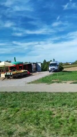 The video shows a roadside stand, possibly selling produce, with multiple umbrellas providing shade. There are stacks of wooden crates, a cart filled with what appears to be corn, and a small structure that might be a food truck or kiosk. A van is parked on the right side of the frame. The scene is set against a backdrop of green fields and a bright blue sky with scattered clouds. The camera appears to be moving, suggesting it's being filmed from a vehicle. The overall impression is that of a rural or agricultural setting on a sunny day.