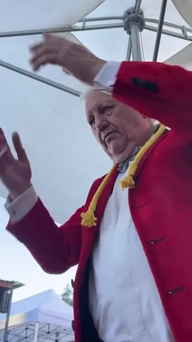 The video shows a close-up of an older white man with gray hair and a white shirt. He is wearing a red jacket and a grey bow tie. A yellow rope with tassels is draped around his neck, resting on his shoulders. The man appears to be speaking, with his mouth open in one frame. His hands are visible, and he is seen adjusting his jacket. He is standing under a white umbrella-like structure. The background shows a tent and some greenery. The camera angle is low, looking up at the man.