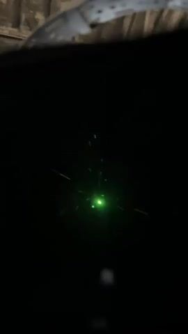 The video captures a close-up view of a black surface with a bright green light emanating from the center. The light appears to be a laser, as evidenced by its concentrated beam and the small, distinct points of light. There are also faint lines or streaks of light, possibly reflections or dust particles illuminated by the laser. The background is dark and out of focus, with a blurry, textured object visible at the top, suggesting an indoor or sheltered environment. The camera is positioned close to the subject, and the overall lighting is dim, emphasizing the brilliance of the green laser. There are no people or other discernible objects in the frame apart from the light source and its effects. The video appears to be a still shot or very slow-moving, focusing on the intensity and color of the laser light.