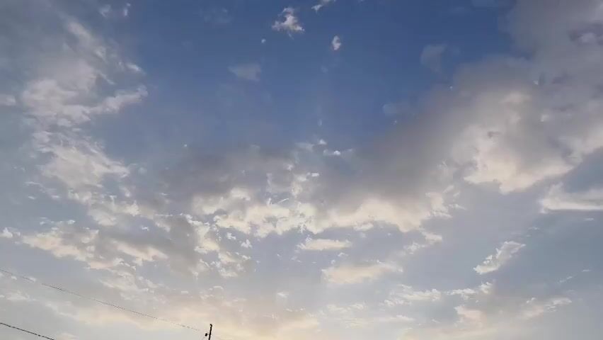 This is a low-angle, static shot of a sky with scattered clouds. The sky is a bright blue, transitioning to a lighter shade towards the bottom of the frame, suggesting the presence of the horizon. The clouds are varied in size and density, with some appearing wispy and others more substantial and textured. Sunlight is visible, filtering through and around the clouds, creating highlights and casting subtle shadows. The overall impression is of a clear day with some cloud cover. There are no discernible people, objects, or actions depicted in the video.