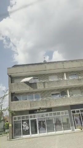 This is a shaky, vertically oriented video shot from inside a vehicle, presumably driving down a street. The sky is bright blue with fluffy white clouds. On the right side of the screen, a multi-story building with light grey brickwork and horizontal windows is visible. A tree with green leaves grows in front of it. A sign on the building's ground floor reads "Revlon" and has a circular sticker with the number "25" on it. To the left, there are several residential buildings with pitched roofs. Further left, a store with a prominent "KIOSK" sign is visible, with a display of ice cream cones. Another building to the left has a sign that reads "Marlboro" and "LOTTO". The street appears to be paved. The video continues to show the building on the right with the "Revlon" sign, then pans to show more buildings and a narrow street between them. Finally, it shows a light grey building with several windows and the number "62" near the entrance.