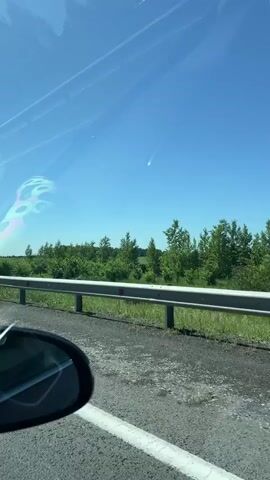 The video, filmed from the perspective of a moving vehicle, shows a clear blue sky with a faint object streaking across it. The object appears to be a white streak with a slightly wider head and a thinner tail, suggesting it could be a comet or meteor. Below the sky, a guardrail runs horizontally across the frame, separating the road from a green, tree-filled landscape. The road itself is visible in the foreground, with a white line marking the edge of the lane. A car's side mirror is also visible in the lower left corner. The lighting suggests it is daytime, with the sun high in the sky.