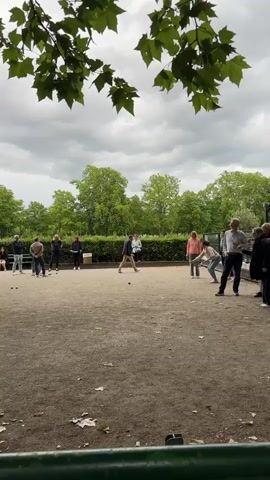 The video captures a group of people playing pétanque in a park. The game is taking place on a gravel court surrounded by greenery and trees. Several individuals are scattered across the court, some in the process of throwing the boules, while others are observing or waiting for their turn. The people are dressed casually in outdoor attire. The sky is overcast, suggesting a cloudy day. The foreground is partially obscured by what appears to be a green structure, possibly a bench or railing. The overall atmosphere is one of leisure and outdoor recreation.