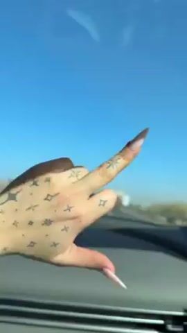 The video shows a hand giving the middle finger while driving down a highway. The hand is fair-skinned with small, dark tattoos of stars and diamonds. The nails are long and pointed with a light pink color. The car is moving at a moderate speed, and other vehicles can be seen in the background. The sky is clear and blue. A Latin song is playing loudly.
