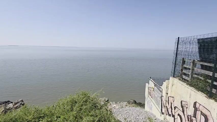 This video shows a wide, calm body of water under a clear, bright sky, suggesting a sunny day. The scene appears to be a coastal area, likely Weston-super-Mare, United Kingdom, given the provided context. In the distance, a long pier extends into the water, with some structures and a crane visible on it. The foreground is characterized by a rocky shoreline with dense green vegetation and some gravel. On the right side of the frame, a concrete wall with graffiti is visible, as well as a section of chain-link fence. The overall atmosphere is serene and quiet, with no people or significant activity depicted, focusing on the natural landscape and man-made structures by the sea. The time of day appears to be daytime, given the bright lighting. There are no discernible emotions conveyed, and the visible text consists of graffiti on the wall, which is not legible in detail.