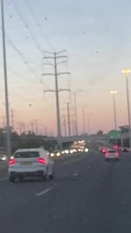The video shows a blurry view from inside a moving vehicle, looking forward. There are cars on the road, with their taillights visible in the distance, and the road appears to be a highway. On the right side of the road, there are streetlights and a bridge or overpass. On the left side, there are tall electrical transmission towers and some trees or vegetation. The sky is a light pink and blue, suggesting either dawn or dusk. The overall image is somewhat out of focus.