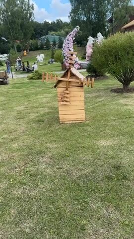 A wooden windmill decoration is positioned in a grassy outdoor area. The windmill is light brown and made of horizontal planks. It has a peaked roof and two rotating blades, each made of several wooden slats. A decorative ladder-like structure is attached to the side of the windmill. In the background, there are people walking, trees, and large floral decorations that appear to be pink and white arches. The ground is covered in green grass.