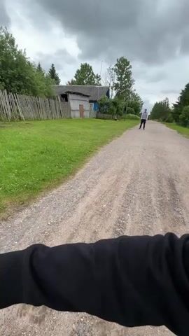 A person walks down a dirt road in a rural setting. The road is lined with grass and trees. There are houses visible on either side of the road. The sky is cloudy. The person is walking away from the camera. The person is wearing a light-colored shirt and dark pants. There is a green object on the ground in the middle of the road.