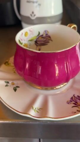 A close-up shot shows a pink and white teacup and saucer with floral designs. The teacup has a gold handle and a detailed floral pattern on the inside. In the background, a white electric kettle with the brand name "delimano" is visible. The objects are placed on a metallic surface, likely a countertop. The lighting appears to be natural, coming from the side and slightly from above, creating subtle shadows.