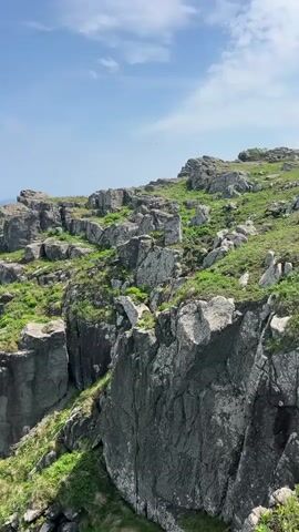 The video shows a rocky cliff face overlooking the ocean. The cliff is composed of large, dark grey rocks, and there is some green vegetation growing between the rocks. The ocean is visible in the background, and the sky is blue with a few clouds. There is a person standing on the edge of the cliff, looking out at the ocean. The camera is positioned at a distance from the cliff, providing a wide view of the scene. The waves are crashing against the base of the cliff. The video has no audio.