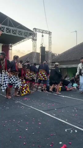 The video shows an outdoor performance with a stage and a crowd of onlookers. Several performers in traditional attire, including checkered patterns and head coverings, are on the ground and in motion. Some are interacting with others who appear to be part of the performance, while others are in a prone position. Flower petals are scattered on the ground. In the background, a stage structure with speakers and lighting equipment is visible. The sky suggests it is dusk.