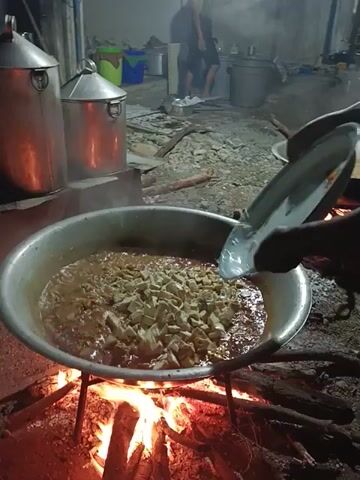 Here's a forensic description of the video:

The video shows a large, oval, metal pot filled with a bubbling, brownish liquid and chunks of what appears to be food. The pot is positioned over a wood fire, with flames visible beneath. The fire is enclosed within a rough, stone-lined area. To the left of the pot, there are several large, stainless steel containers with conical tops. In the background, a person is partially visible, lying down. The overall setting appears to be an outdoor or semi-outdoor kitchen or cooking area, likely for a large event or gathering. The video is shot from a slightly elevated perspective, looking down into the pot.
