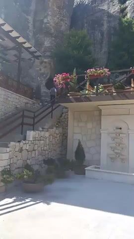A sunny day scene shows the exterior of a building situated against a massive rock face. A wooden balcony, adorned with potted pink flowers and greenery, extends across the upper level of the building. Below the balcony, a white stone wall features a multi-tiered water fountain with cascading water. Several small, conical evergreen trees in pots are placed on the ground in front of the wall. To the left, a stone staircase leads up to the balcony area, with a railing and more potted plants. People are visible on the staircase, ascending or descending. The background is dominated by the imposing, textured surface of the rock cliff.