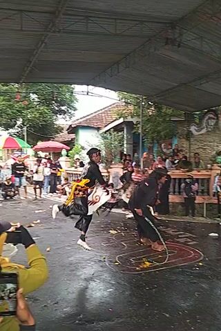 The video shows a cultural performance taking place in an outdoor setting under a corrugated metal roof. There's a crowd of people watching. Two individuals dressed in black traditional attire are performing movements that appear to be part of a ritualistic dance. They are barefoot on a wet surface. The performance space has a design marked on the ground with what appears to be colored powder. There are also decorative masks or headdresses among the audience, which seem to be part of the cultural display. The area is surrounded by greenery and what looks like residential buildings in the background.