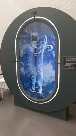 This media file shows an interactive exhibit featuring a life-size display of a cosmonaut in a spacesuit. The cosmonaut is depicted floating against a backdrop of clouds and the Earth, creating the illusion of being in space. The display is housed within a large, dark gray oval frame with a glowing white interior ring. A red arrow on the screen points downwards with the text "встаньте на круг" (stand on the circle), suggesting an interactive element where viewers are meant to position themselves on a marked area.

As the video progresses, the display changes. A circular gauge with the number "10" appears around the cosmonaut's head, and circular targeting reticles are superimposed on the image. The spacesuit is white with various pouches and tubes. The helmet's visor reflects a dark interior, and the inscription "СССР" (USSR) is visible on the helmet. The overall scene appears to be indoors, likely in a museum or exhibition hall, given the lighting and the presence of a white chair and a dark gray base supporting the display. The context "Saburovka, Russia" suggests this exhibit is located in that region. No specific time of day or weather conditions are discernible as the setting is indoors. The tone of the exhibit is educational and possibly celebratory of space exploration.