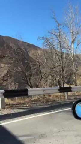 The video shows a car driving along a road with a white and black guardrail.  The landscape includes trees, some with yellow leaves, and a large mountain range in the background under a clear blue sky. The camera seems to be positioned inside the car, capturing the view out the side window. A triangular road sign is briefly visible. The overall impression is of a scenic drive through a mountainous area.