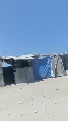 A video shows a row of makeshift structures built from corrugated metal and tarps in a sandy, outdoor environment under a clear blue sky. The structures appear to be temporary shelters, with some showing signs of wear and tear, such as torn tarps and rusted metal. The ground is covered in sand and scattered debris. The camera pans across the structures, providing a view of their construction and the overall setting. No people are visible in the video.