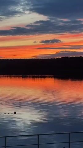 A video captures a sunset over a body of water. The sky is filled with vibrant colors of orange, pink, and blue, with clouds streaking across. The sunset is reflected in the calm water, creating a mirror image of the sky. In the foreground, a wooden deck with a railing and some outdoor furniture is visible, suggesting a scenic viewpoint for observing the sunset. A dark, silhouetted treeline borders the water in the distance. The overall mood is peaceful and serene.