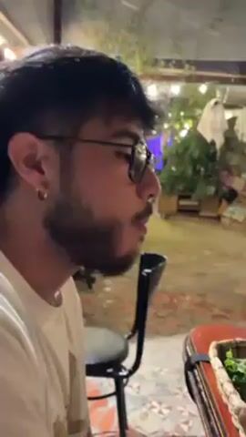The video shows a group of people sitting at a wooden table outside in an outdoor dining setting. There are various baskets on the table containing food, including lettuce, bread, and fried sticks. A man wearing glasses and a beard is seen speaking in the first shot. Other people are visible at the table, one of whom is eating a sandwich. There is also a pizza on a wooden board. The table is cluttered with items like a phone, a red-checked cloth, a wine glass, a beer bottle, and a deck of Uno cards. String lights hang in the background, adding to the outdoor ambiance.