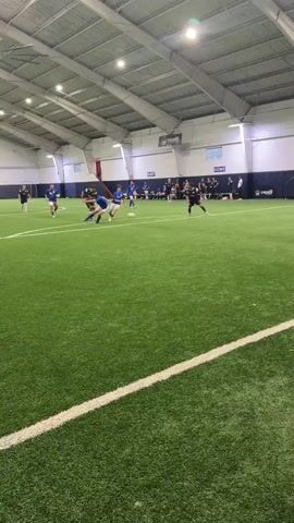The video shows a soccer game taking place indoors on an artificial turf field. The field has white lines marking boundaries and divisions. A soccer goal with netting is visible on the right side of the frame. Several players are on the field, some in blue jerseys and others in black or dark jerseys. One player in a yellow jersey and gloves, presumably the goalkeeper, is positioned near the goal. The ceiling of the indoor facility is visible, with exposed beams and lighting fixtures. Spectators can be seen in the background, seated in bleachers. The players appear to be engaged in a game, with some actively kicking or chasing the ball. The action suggests a dynamic play, with players moving and interacting on the field. The overall environment is that of a sports arena.