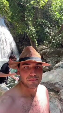 A close-up selfie video captures a young man wearing a light brown fedora with a black band. He has short dark hair, brown eyes, and is shirtless, revealing a gold necklace with a pendant. He is in a natural setting, likely a forest or wooded area, with lush green foliage overhead and a waterfall cascading into a clear blue pool of water in the background. Sunlight filters through the trees, casting dappled shadows on the man's face and shoulders. The water appears cool and inviting. Another person, partially visible in the background, is also in the water. The overall mood is relaxed and adventurous, suggesting a summer day spent exploring nature. The time of day appears to be daytime, given the sunlight. There is no visible text in the media file.